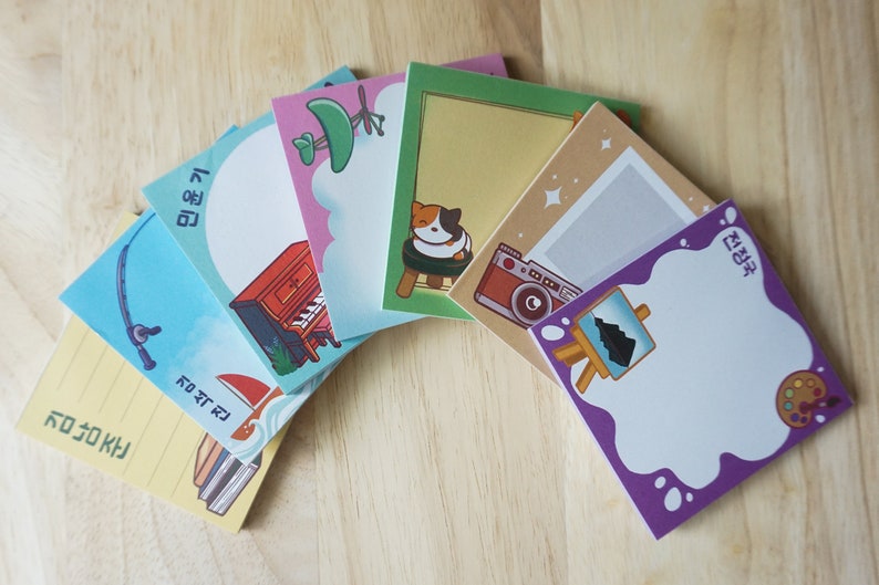 May include: A selection of square sticky notes featuring illustrations and Korean text. Designs include a fishing rod, piano, cat, camera, and paint palette. The notes are arranged on a light wood surface.