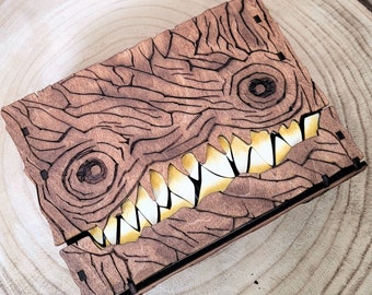 May include: A wooden box with a carved monster face design. The monster has two large eyes and a wide, toothy grin. The box is made of dark brown wood and has a natural wood grain.