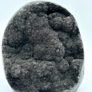May include: A dark gray geode with a rough, textured interior. The geode has a rounded shape with a smooth, polished exterior that transitions to a teal and gray color near the base. The interior is filled with a cluster of dark gray crystals.