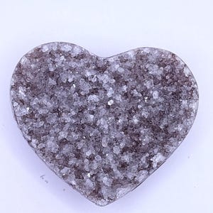 May include: A heart-shaped, dark gray geode with a sparkling, crystalline surface. The geode has a rough, textured appearance and is set against a white background. The heart shape is clearly defined, making it a unique decorative item.