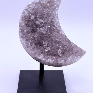 May include: A crescent-shaped amethyst crystal geode, mounted on a black metal stand. The geode is a light purple color and has a crystalline texture. The stand has a square base and a thin, vertical support.