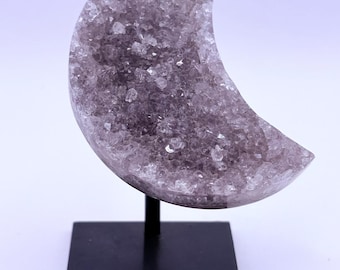 May include: A crescent-shaped amethyst crystal geode, mounted on a black metal stand. The geode is a light purple colour and has a crystalline texture. The stand has a square base and a thin, vertical support.