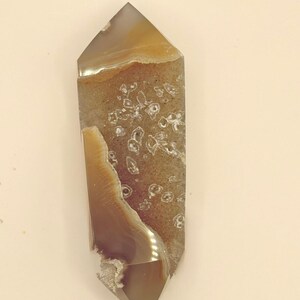 May include: A polished agate crystal point with a hexagonal shape. The crystal features bands of brown, tan, and white, with a translucent quality. The stone has a natural, organic appearance, suitable for display or collection.