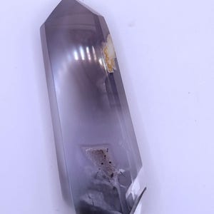 Pode incluir: Um grande cristal de quartzo fumê polido com uma ponta. O cristal é de cor cinza translúcido com inclusões esfumaçadas mais escuras e algumas áreas de descoloração. O cristal é exibido em um fundo branco.