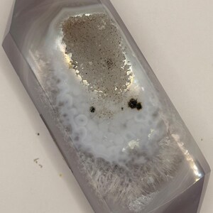May include: A polished, double-terminated agate crystal in shades of gray and white. The crystal features a central geode cavity with sparkling, granular inclusions. The exterior has a smooth, faceted surface, showcasing natural banding patterns.