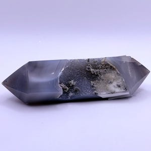 May include: A double-terminated, polished crystal with a gray and white color scheme. The crystal has a central cavity with a rough, textured appearance. The overall shape is a long, hexagonal prism, ideal for display or collection.