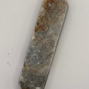 Pode incluir: Uma ponta de cristal de dupla terminação com uma superfície áspera e texturizada. O cristal apresenta uma mistura de tons cinza, marrom e branco, com estruturas cristalinas visíveis. A forma é um longo prisma retangular com extremidades pontiagudas.