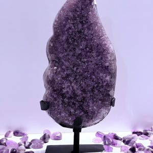 Könnte beinhalten: Eine große, polierte Amethyst-Geode in Lilatönen wird auf einem schwarzen Metallständer präsentiert. Die Geode hat eine kristalline Struktur und ist von kleineren Amethyststeinen umgeben. Der Hintergrund ist ein weiches, helles Lila.