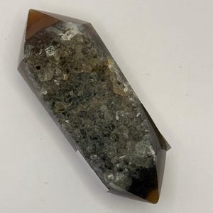 Pode incluir: Cristal de dupla terminação com ponta castanho escuro e corpo transparente e texturizado. O cristal tem formato retangular com extremidades pontiagudas. O interior é preenchido com pequenas formações cristalinas claras.
