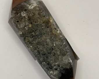 Pode incluir: Cristal de dupla terminação com ponta castanho escuro e corpo transparente e texturizado. O cristal tem formato retangular com extremidades pontiagudas. O interior é preenchido com pequenas formações cristalinas claras.