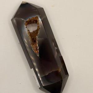May include: A dark gray crystal point with a hollowed-out center revealing a brown and orange interior. The crystal has a polished, faceted exterior and a natural, rough interior. The crystal is a gemstone.