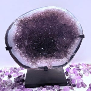 May include: A large, polished amethyst geode, showcasing a deep purple crystal interior. The geode is mounted on a black stand, surrounded by smaller amethyst stones. The outer edge of the geode is a dark gray.