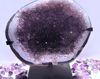 Pode incluir: Uma grande geodo de ametista polida, mostrando um interior de cristal roxo escuro. A geodo é montada em um suporte preto, rodeada por pedras de ametista menores. A borda externa da geodo é cinza escuro.