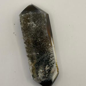 May include: A polished crystal point with a dark brown tip and base, transitioning to a gray and white speckled center. The crystal has a faceted shape and a natural, earthy appearance.