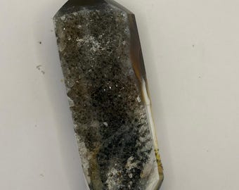 Peut inclure: Une pointe de cristal polie avec une pointe et une base brun foncé, passant à un centre tacheté de gris et de blanc. Le cristal a une forme à facettes et une apparence naturelle et terreuse.