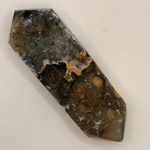 May include: A polished, elongated crystal with a faceted top and bottom. The crystal features a mix of colors, including brown, gray, black, and hints of orange. The surface has a textured appearance, with visible crystalline structures.
