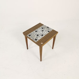May include: A small, square wooden side table with a mosaic tile top featuring a pattern of black, gray, and white tiles. The table has four tapered legs and a dark wood frame. A small circular emblem is visible on the side.