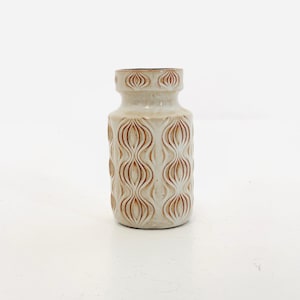 Könnte beinhalten: Eine beige Keramikvase mit einem sich wiederholenden, wellenförmigen Muster in Braun. Die Vase hat einen schmalen Hals und einen breiteren Boden. Der obere Teil der Vase hat kleine, ovale Vertiefungen. Ein dekorativer Artikel.