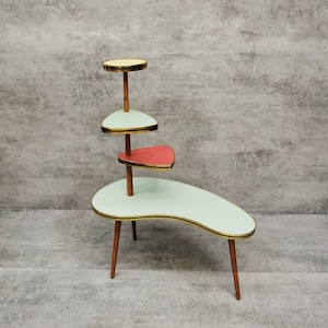 May include: A vintage plant stand with four tiers in pastel colors. The stand has a kidney-shaped base, a triangular shelf, and two circular shelves. The legs are wooden, and the edges are trimmed in gold.