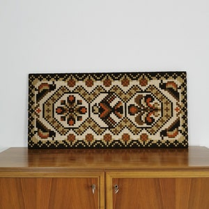 Könnte beinhalten: Ein rechteckiger, gewebter Wandbehang mit einem geometrischen Muster in Braun-, Beige- und Schwarztönen. Das Design zeigt florale und geometrische Formen. Der Wandbehang wird über einem Holzschrank ausgestellt.