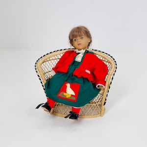 Puede incluir: Una muñeca vintage con cabello castaño y ojos azules, vestida con una chaqueta roja, un vestido verde con una aplicación de pato, calcetines rojos y zapatos negros. La muñeca está sentada en una silla de mimbre con ribete en blanco y negro.