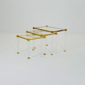 Könnte beinhalten: Satz von drei Beistelltischen. Jeder Tisch hat eine transparente Acrylbasis mit goldfarbenen Metallakzenten. Die Tischplatte ist aus klarem Glas. Die Tische sind in einer ineinander verschachtelten Konfiguration angeordnet.