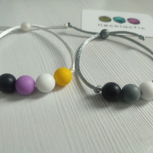 Könnte beinhalten: Zwei Armbänder mit unterschiedlich farbigen Perlen. Das erste Armband hat ein weißes Band und schwarze, lila, weiße und gelbe Perlen. Das zweite Armband hat ein graues Band und schwarze, graue, weiße und burgunderrote Perlen. Die Armbänder liegen auf einer weißen Oberfläche mit einer Karte, auf der "necklactic" steht.