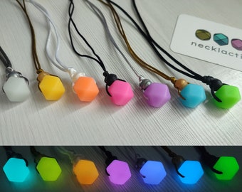 May include: Eight glow-in-the-dark necklaces with different colored geometric pendants. The necklaces are displayed on a white surface. The pendants are in the following colors: white, yellow, orange, pink, purple, blue, green, and lime green. The necklaces are made by necklactic.