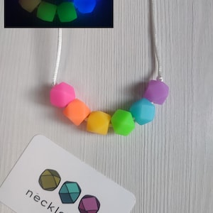 Peut inclure: Collier en silicone arc-en-ciel avec des perles qui brillent dans le noir. Le collier est fait d'un cordon blanc et comporte sept perles dans un motif arc-en-ciel. Les perles sont en forme de pentagone. Le collier est présenté sur une surface en bois clair. L'image montre également un gros plan des perles qui brillent dans le noir.