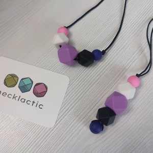 May include: Two necklaces with colourful silicone beads. One necklace has purple, black, white, and pink beads. The other necklace has purple, black, blue, and white beads. The necklaces are on a white wooden surface. The text "necklactic" is on a white card.