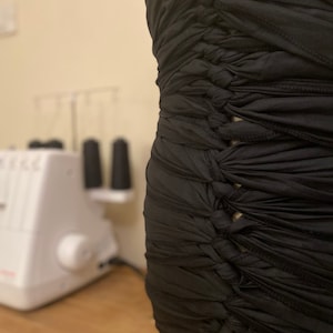 Puede incluir: Un vestido negro sin tirantes con un diseño texturizado único. La tela está fruncida y anudada, creando un efecto de capas. El vestido se exhibe en un maniquí, mostrando su forma y detalles. En el fondo se ven una máquina de coser y carretes de hilo.