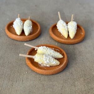 Pode incluir: Três pequenos pratos de terracota, cada um contendo duas réplicas em miniatura de espigas de milho em palitos de madeira. O milho é amarelo com um revestimento branco texturizado que lembra queijo ou tempero, disposto nos pratos de barro.