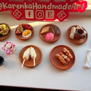 Pode incluir: Uma coleção de alimentos em miniatura de argila em pequenos pratos de terracota, incluindo tacos, ovos, pão, churros e milho. Os acessórios incluem um pilão de madeira, uma tigela preta e duas vassouras em miniatura. Placa vermelha @KarenkaHandmadeArt.
