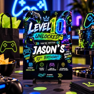 May include: A rectangular birthday invitation card with a black background featuring neon green, blue, and yellow gaming graphics. The text reads Level 10 Unlocked, Jason's 10th Birthday, and includes event details like date, time, location, and RSVP contact.
