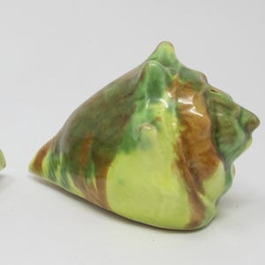 May include: Two ceramic seashell figurines. The shells are painted with a glossy finish in shades of brown, green, and yellow. The figurines are triangular in shape and sit on a white surface.
