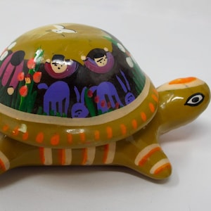 May include: Hand-painted ceramic turtle figurine in shades of olive green, orange, and cream. The turtle's shell features a colorful scene with figures, flowers, and rabbits. The figurine is approximately 6 inches long.