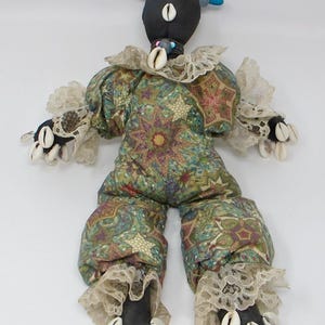 May include: A handmade doll with a black head adorned with blue beads and cowrie shells. The doll wears a patterned outfit with lace trim and more cowrie shells on the hands and feet. The doll is approximately 18 inches tall.