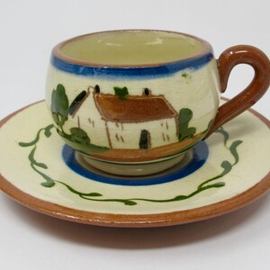May include: A ceramic cup and saucer set featuring a hand-painted cottage scene. The cream-colored cup has a brown handle, a blue rim, and a brown-roofed house illustration. The matching saucer has a blue center ring and green vine accents.