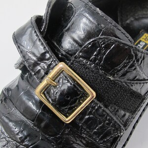 May include: Black leather shoes with a crocodile pattern and a gold buckle. The shoes have a strap across the top and a black elastic band.