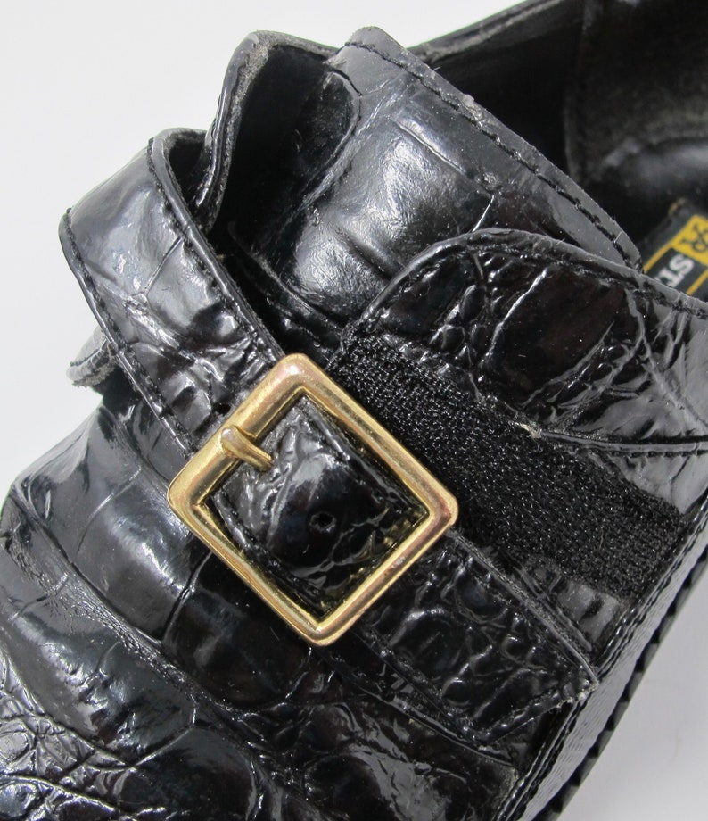 May include: Black leather shoes with a crocodile pattern and a gold buckle. The shoes have a strap across the top and a black elastic band.
