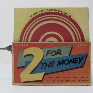 May include: A vintage red, white, and blue cardboard box with a timer inside. The box has the words "2 for the money" printed on it. The timer is a circular disc with a red center and white rings. The box has a lever on the side that is used to start the timer.