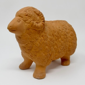May include: A terracotta-colored clay figurine of a ram standing in profile. The sculpture features a textured, curly wool pattern covering the body, a smooth face, and curved horns on the side of the head. The ram is depicted with four legs.