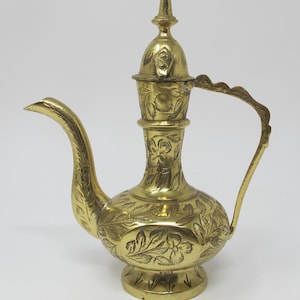 May include: A gold-colored, ornate brass pitcher with a long spout and a curved handle. The pitcher features intricate floral and leaf engravings across its body, lid, and handle. The base is round and slightly flared, and the lid is topped with a small finial.