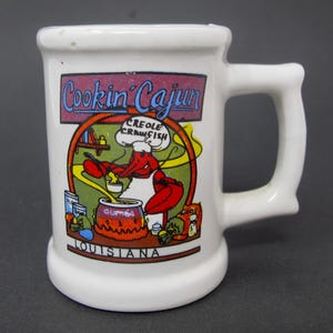 May include: A white ceramic mug featuring a colorful graphic of a red crawfish wearing a chef hat and stirring a pot labeled Gumbo. The text Cookin' Cajun appears at the top, and Louisiana is printed at the bottom of the illustration.