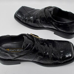 May include: A pair of black leather shoes with a buckle closure. The shoes have a crocodile-embossed pattern and a square toe. The brand name "Stacy Adams" is visible on the inside of the shoe.