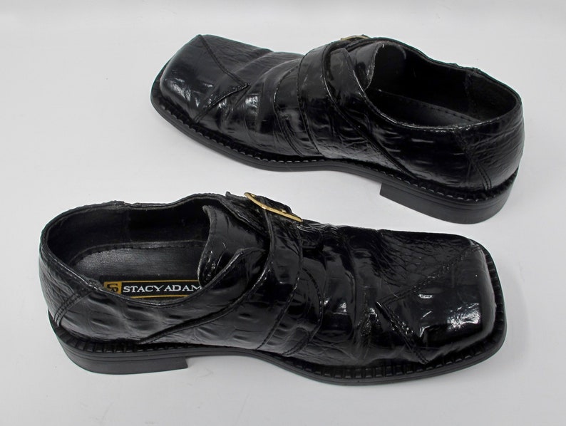 May include: A pair of black leather shoes with a buckle closure. The shoes have a crocodile-embossed pattern and a square toe. The brand name "Stacy Adams" is visible on the inside of the shoe.
