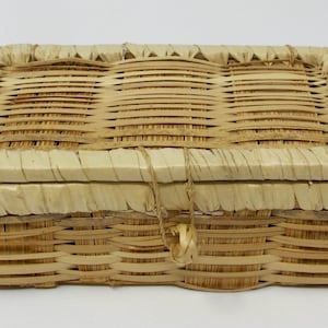 May include: A rectangular woven basket with a lid. The basket is made of light brown woven material. The lid is attached with a string. The basket is likely used for storage or decoration.