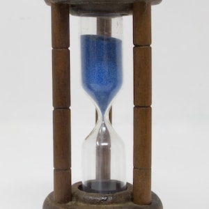 May include: A wooden hourglass with a clear glass center filled with blue sand. The hourglass has a dark brown wooden frame and base. The design is simple and classic, suitable for decorative purposes or timing.