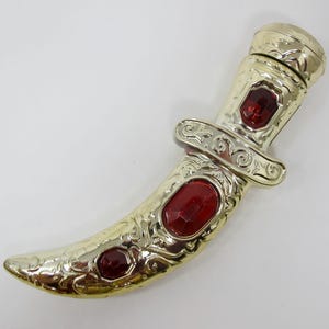 May include: Gold-colored decorative bottle with a dagger shape and red glass gems. The bottle has an ornate design.