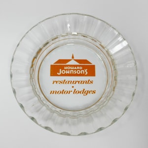 May include: A white glass ashtray with a gold rim and an orange logo that reads "Howard Johnson's restaurants motor lodges".